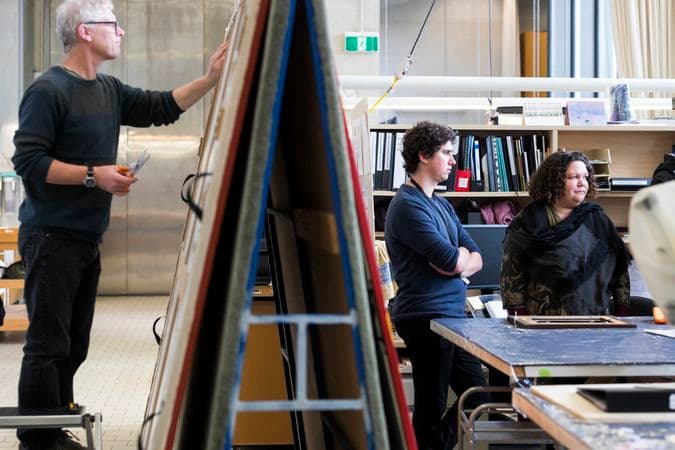 In a conservation lab one man is standing on a footstool and treating a paining resting on an a-frame stand. Nearby, two other people are watching a demonstration out of frame..