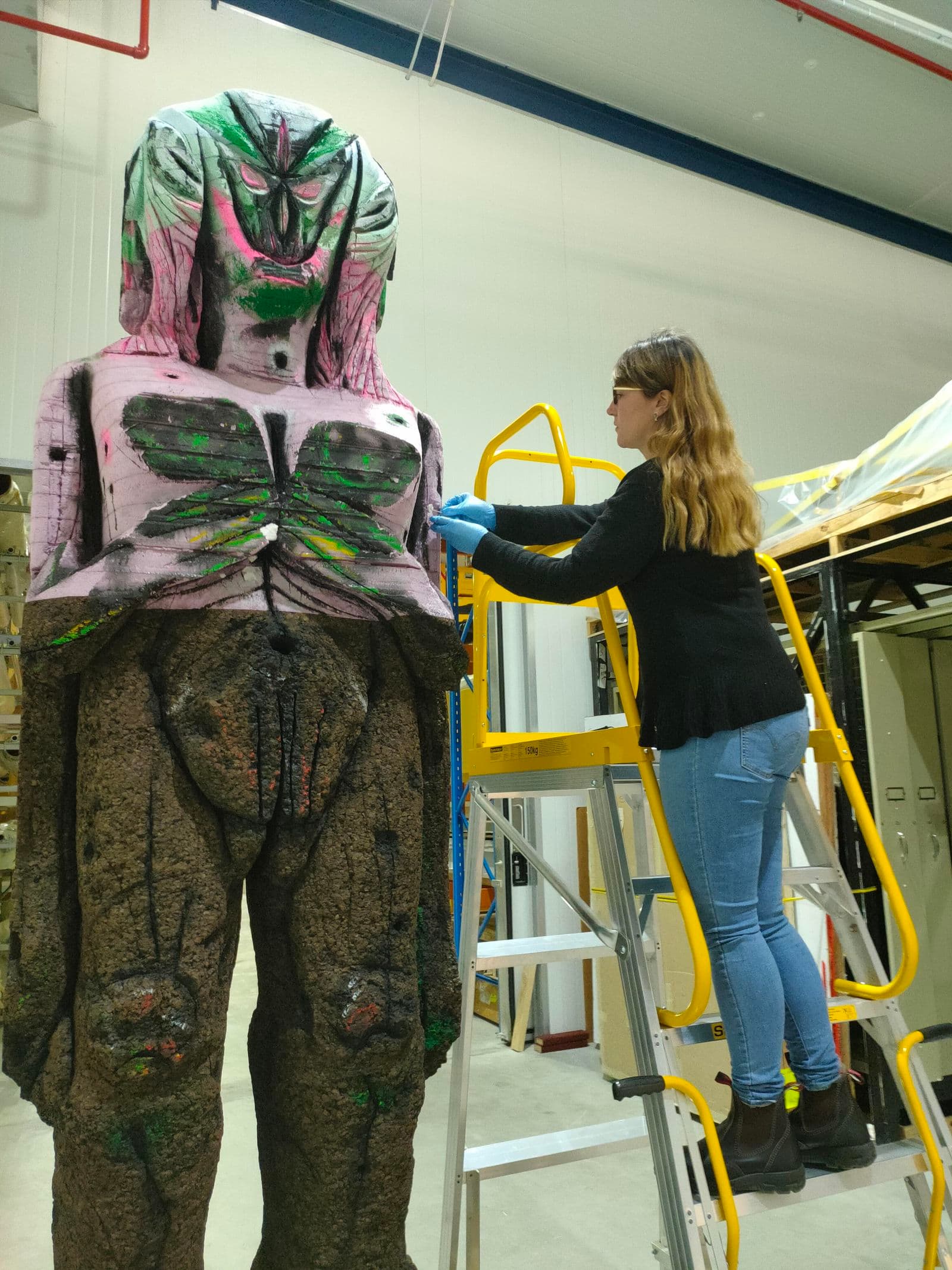 Photograph of Objects Conservator, Kim Goldsmith, standing on a ladder, wearing gloves treating a large oversized sculpture by contemporary artist Huma Bhabha Waiting for another game