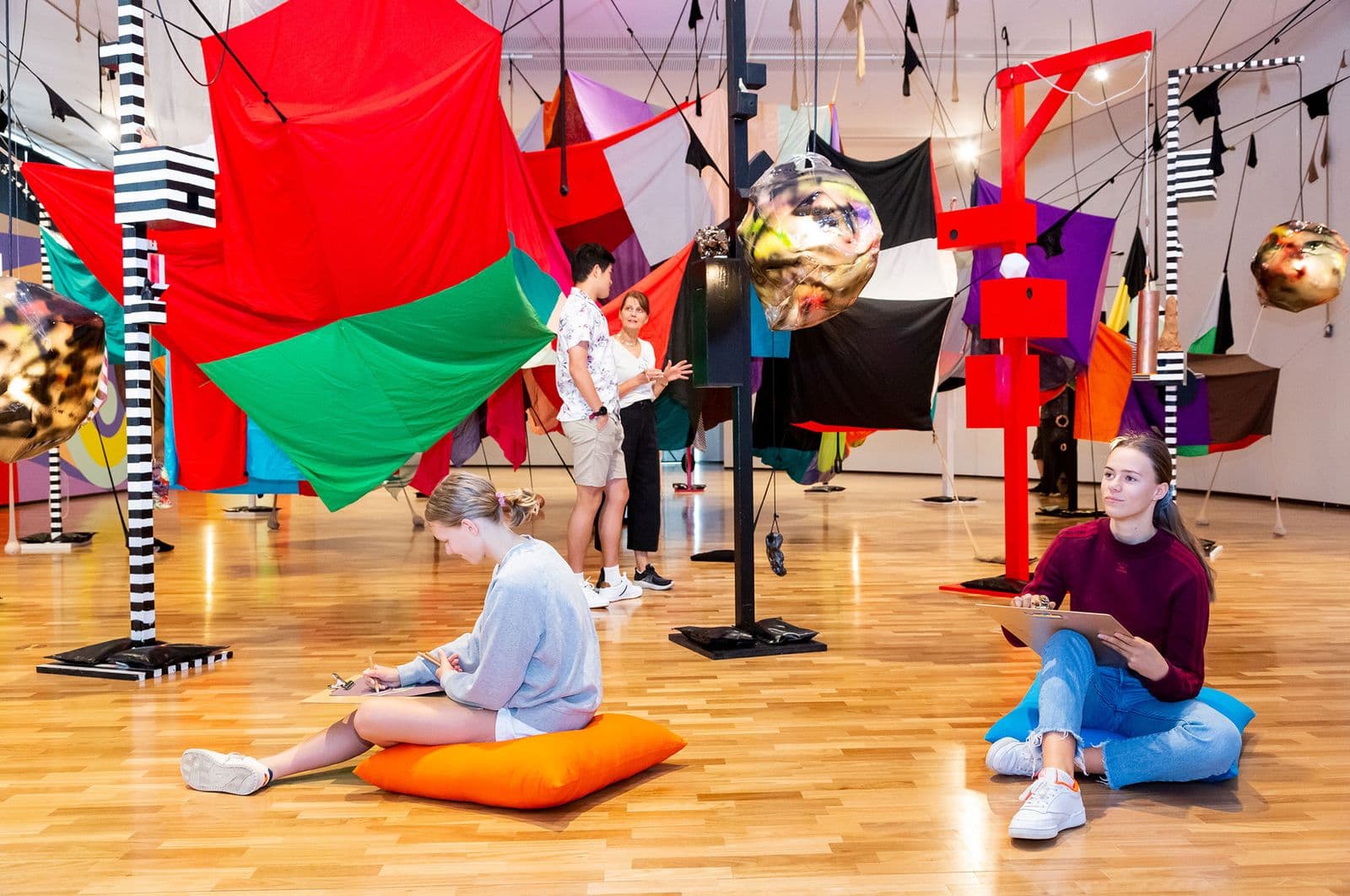Two people sit and draw while two others stand within a large full-room installation made of brightly coloured fabric and poles