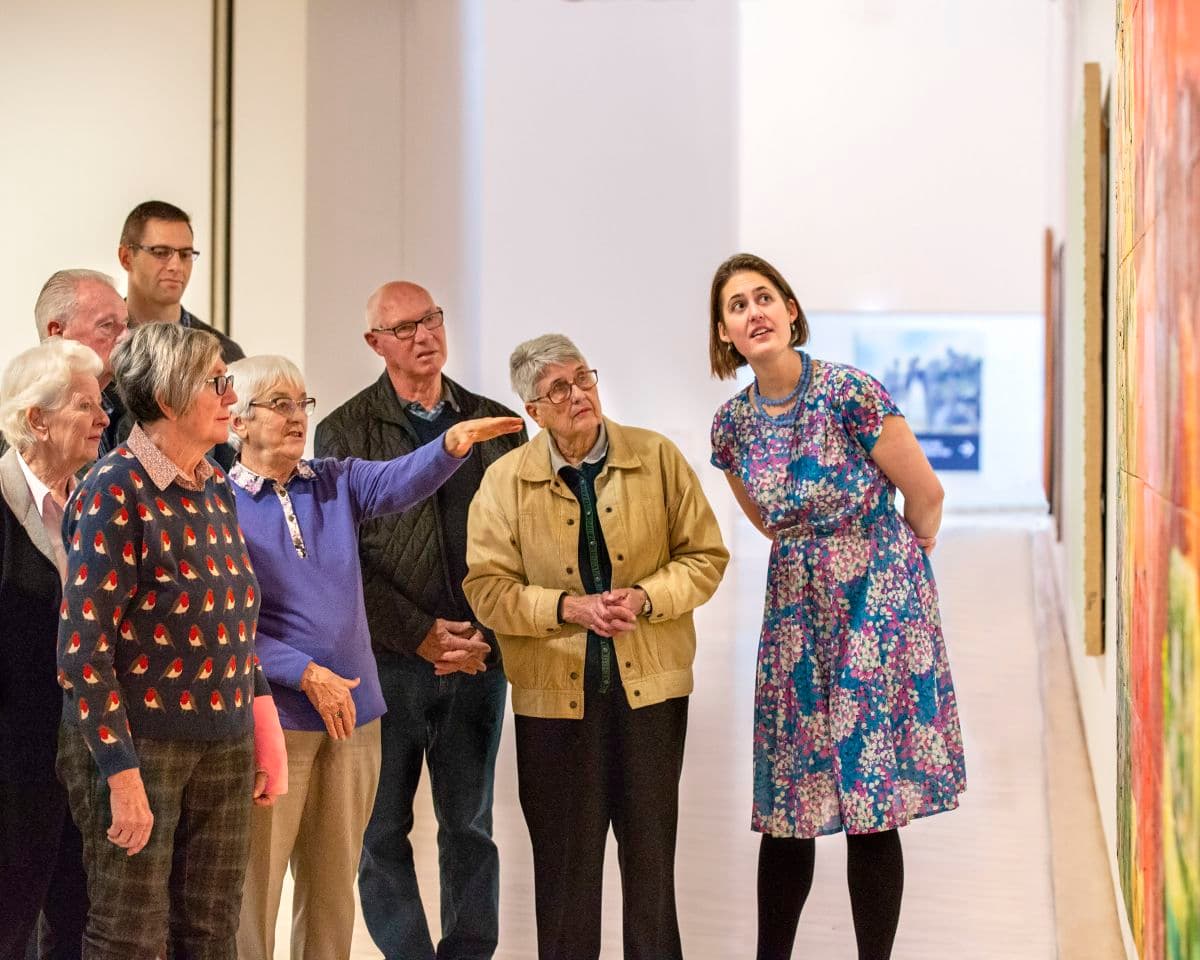 A group of people stand in front of a painting closely examining and talking about it
