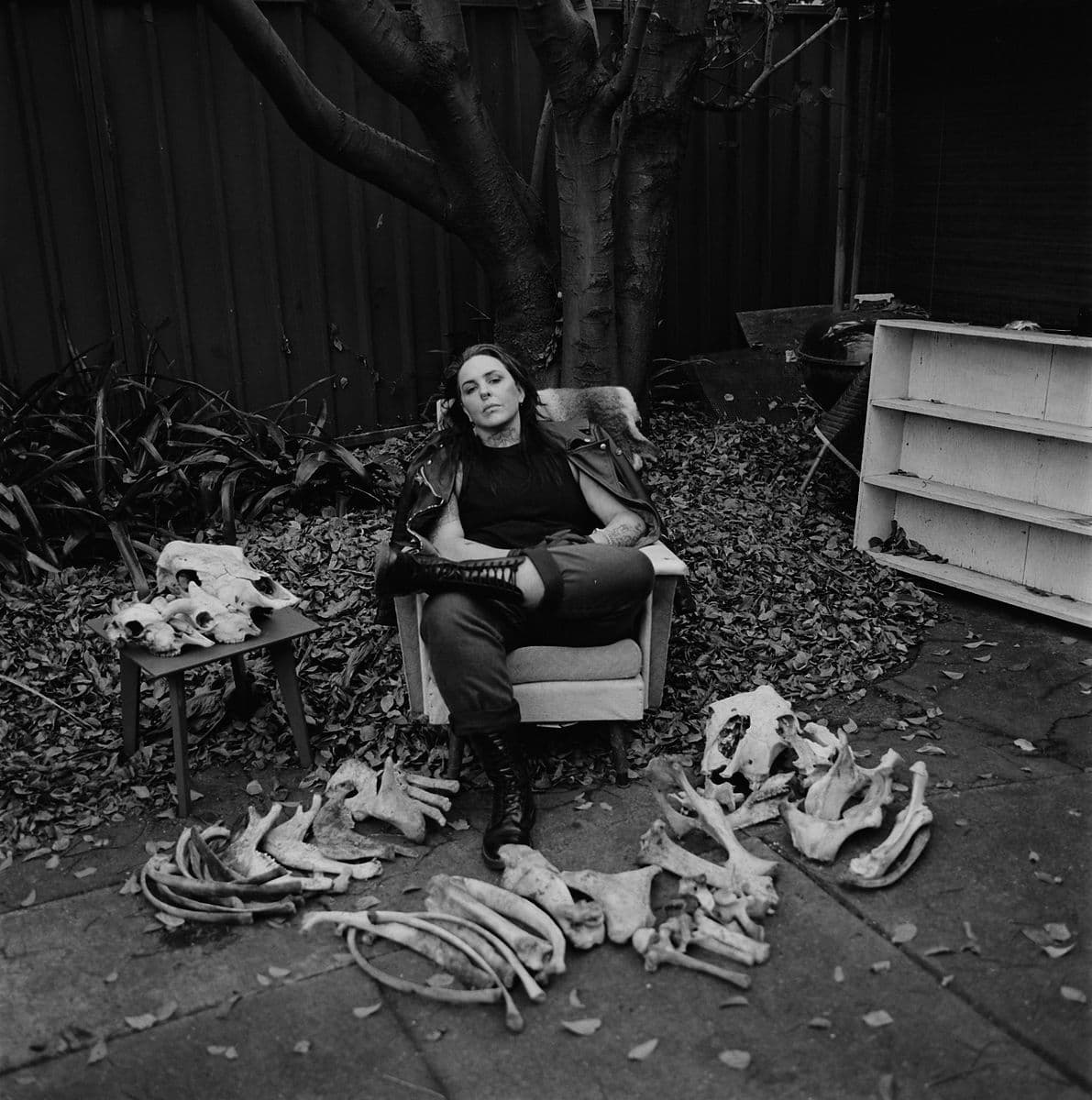 Black and white photographic portrait of SJ Norman sitting in a chair, surrounded by dry leaves and animal bones.