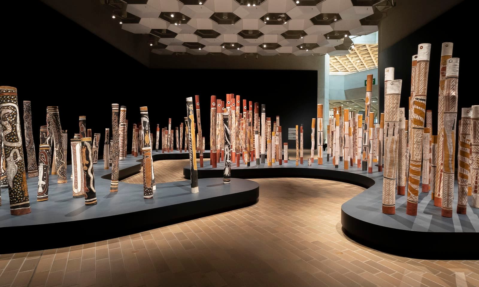 A wide-angle photograph showing an installation of 200 painted hollow logs standing upright in a large gallery space. The surrounding walls are painted in a dark colour and the logs are positioned on a navy plinth shaped to resemble both sides of a river bank.