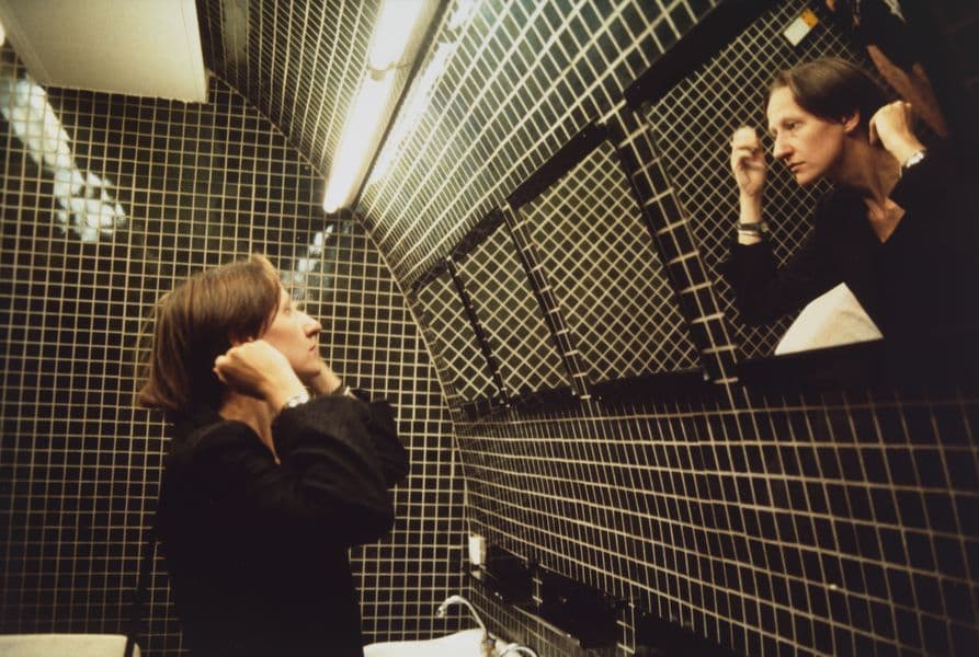 Friend of artist Nan Goldin staring into a mirror in a green tiled bathroom.