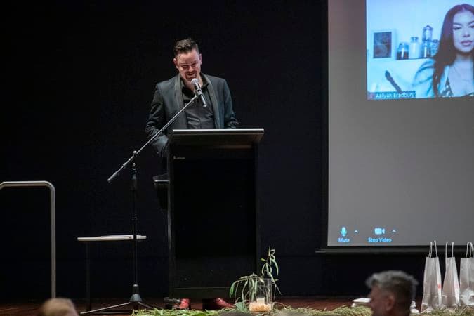 A man is standing behind a lectern on a stage, smiling as he give a speech. There is a projection of people joining via video call displayed on the stage beside him.
