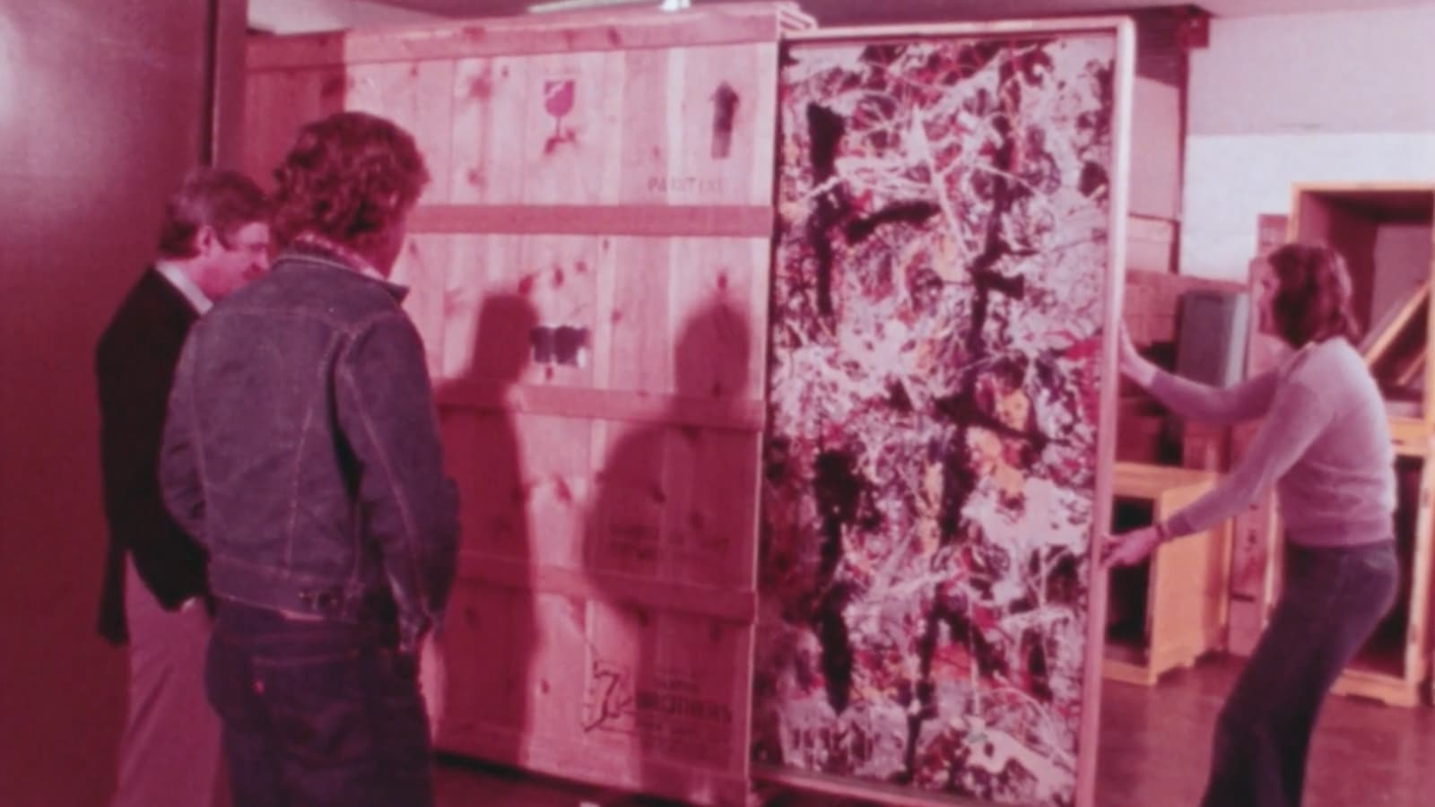 A video still of one man pulling a large painting out of a crate while two other men watch