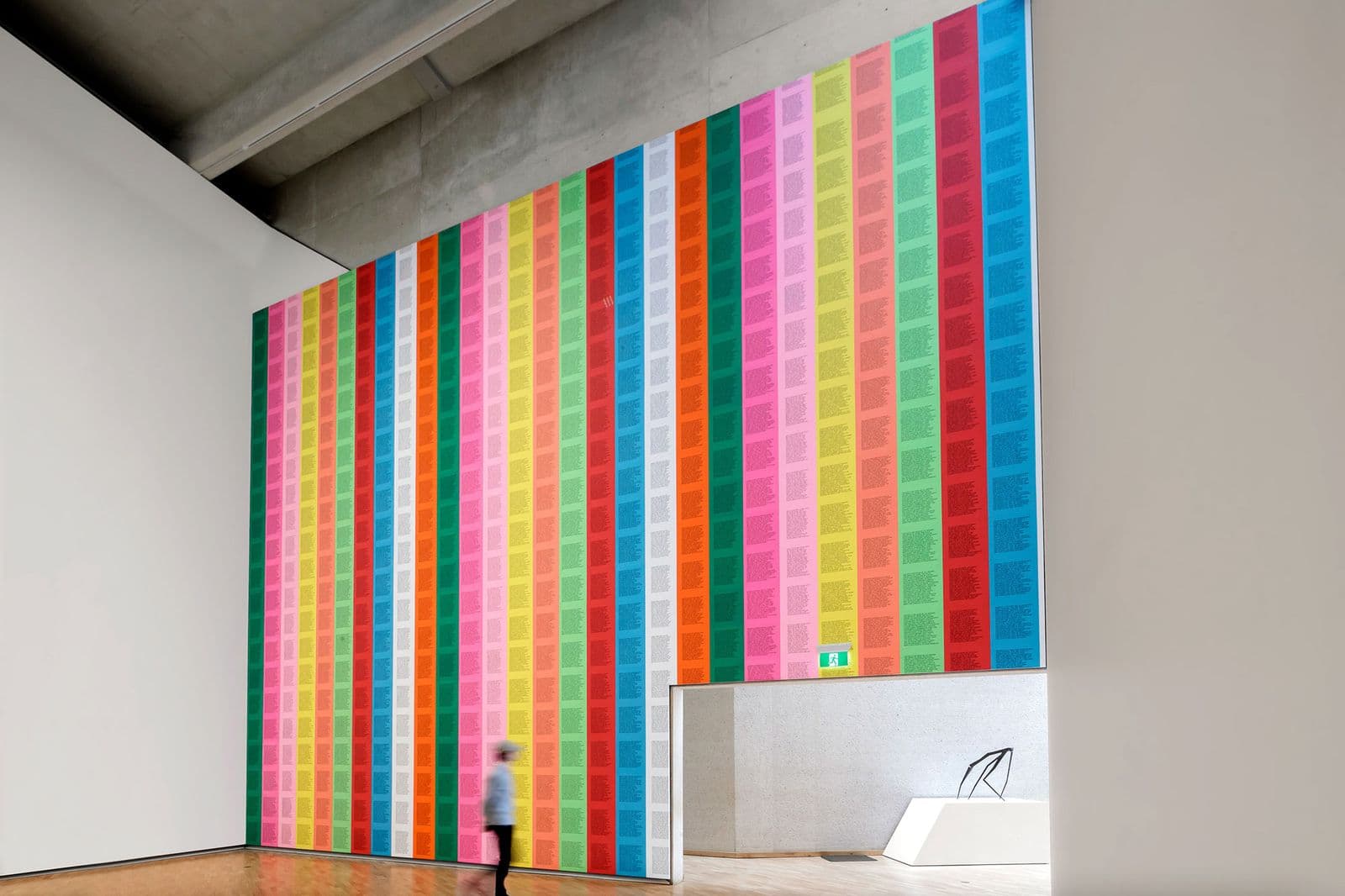 In a high-ceilinged gallery space a woman walks near a wall pasted completely with coloured squares of writing