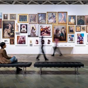 A woman is sitting in font of a large wall of many portraits in a large gallery space