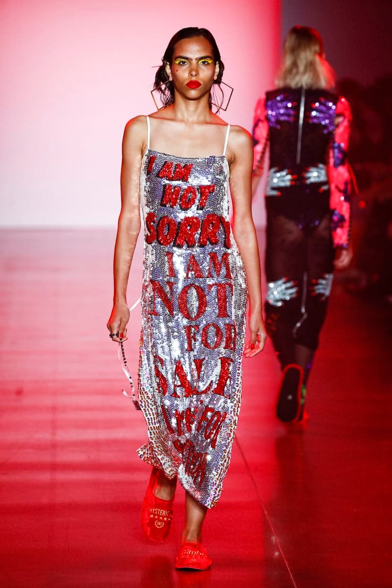 Photograph of a model on a catwalk wearing an embellished slip dress with writing on the front saying 'I am not sorry, I am not for sale, I am not for reproduction'