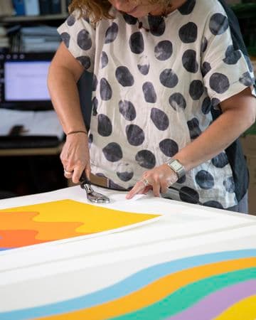 A conservator stands at table working on two brightly-coloured works of art
