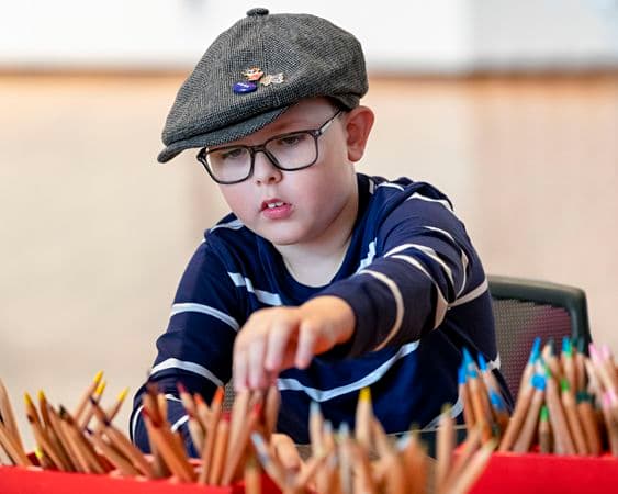 Young boy wearing glasses and a cap reaching for coloured drawing pencils