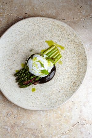 A plate with a poached egg placed on top of five asparagus stalks with a dark, silky sauce at the base