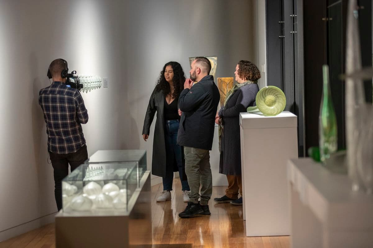 Three people are standing in a gallery looking at works of art while one person with a video camera films the art