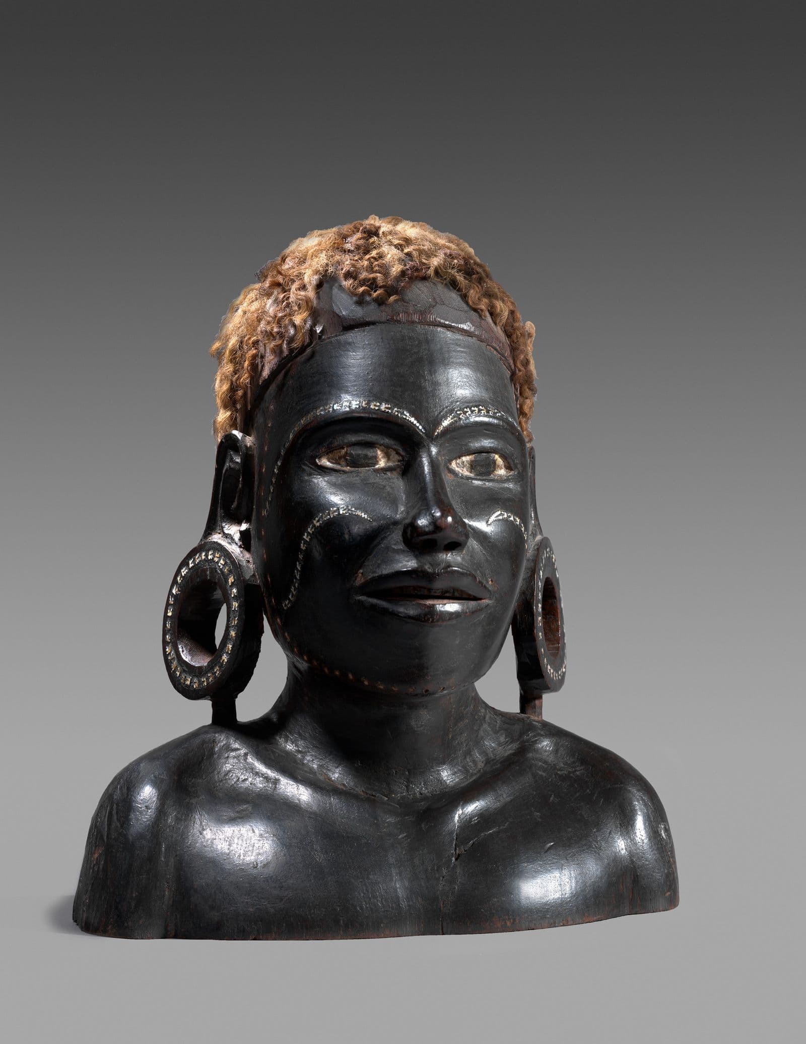 Bust of a young man with stretched earlobes and light curly hair. There are decorative markings on the face and earlobes