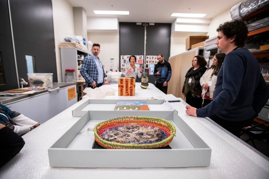 Indigenous art objects are sitting on a large table in a conservation lab. People are standing around the table, one woman is speaking to the group.
