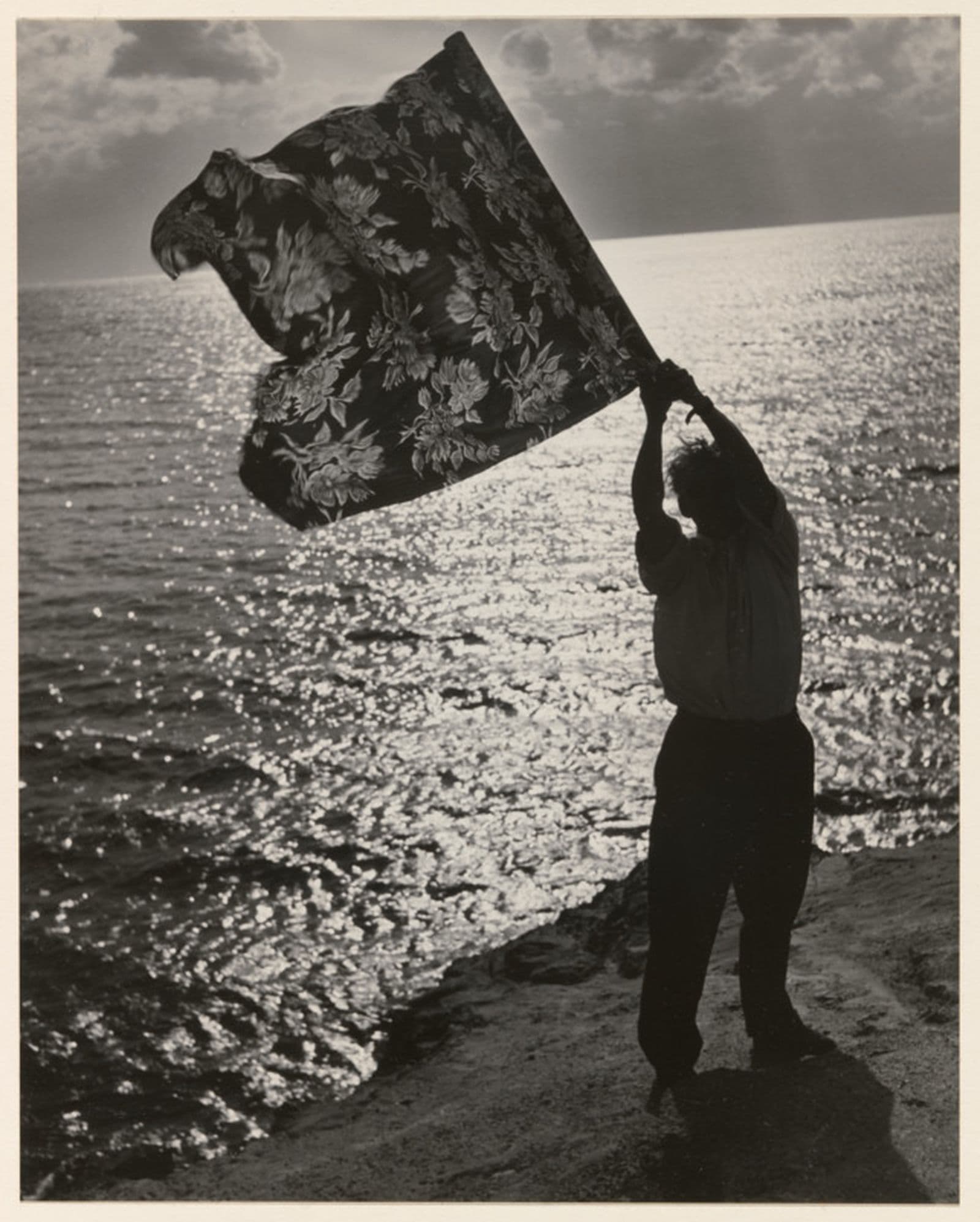 A black and white photograph of a figure standing at the edge of the cliff holding a piece of fabric that flutters in the wind