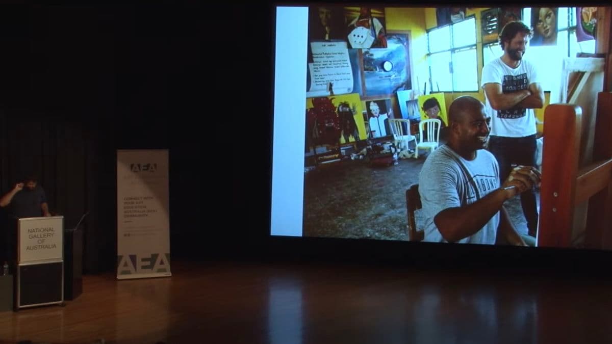 Video still of man giving presentation with slideshow playing alongside.