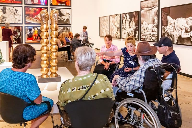 a group of people, some in wheelchairs, sit around a sculpture listening to a guide