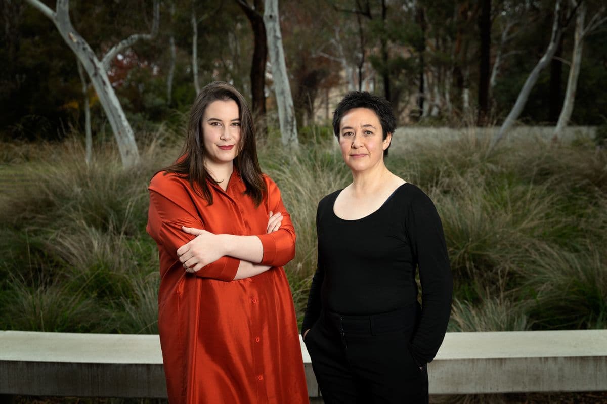 Two smiling women photographed in front of greenery, one with long brown hair dressed in red (AJ America) and the other with short black hair dressed in black (Sally Whitwell)