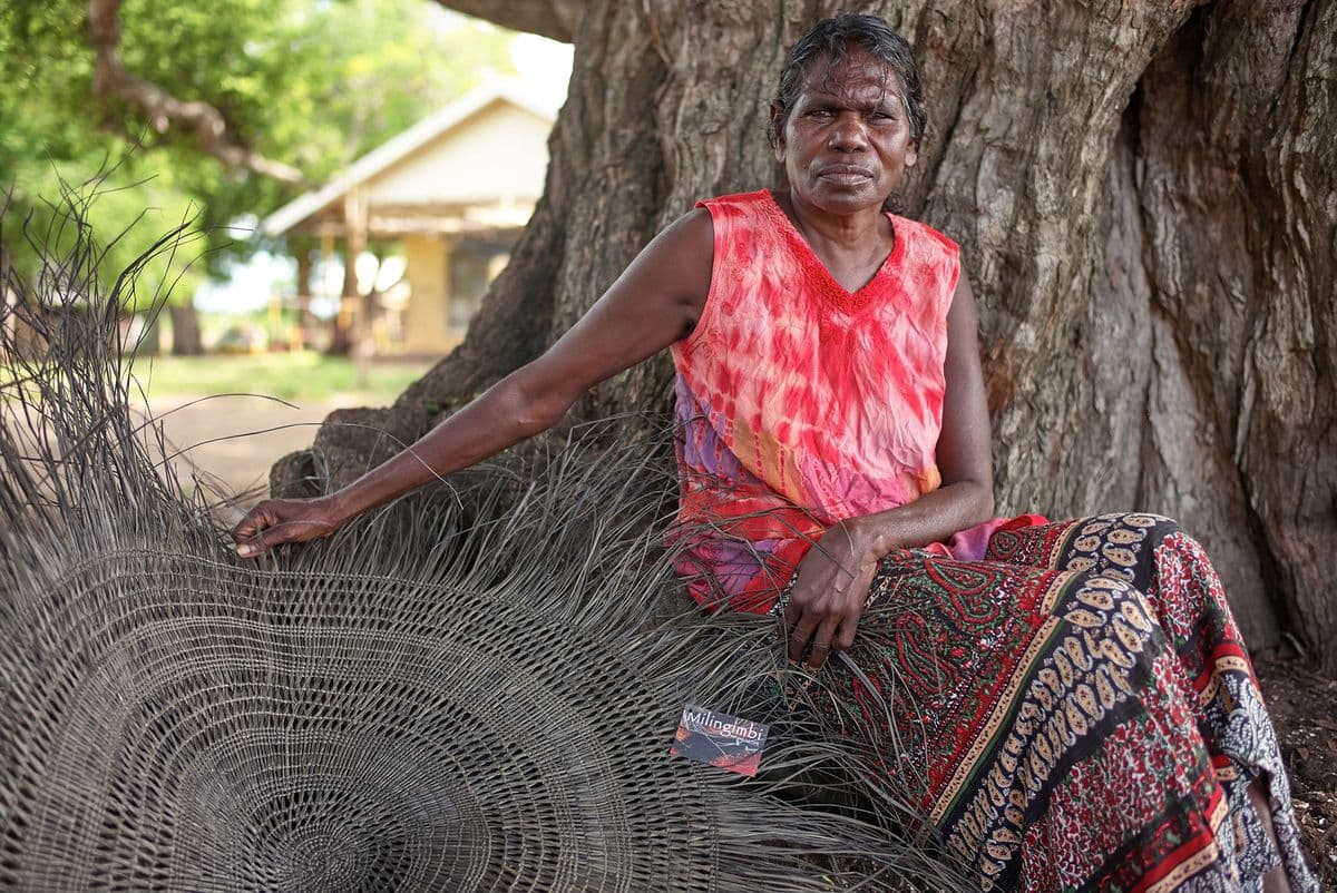 Helen Ganalmirriwuy sits beneath a tree with her woven artwork