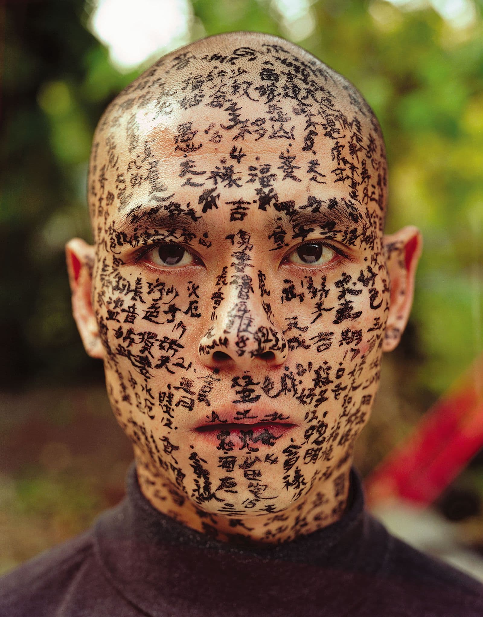 Close-up photograph of a man with his face, neck, ears and scalp covered in Asian characters in black ink
