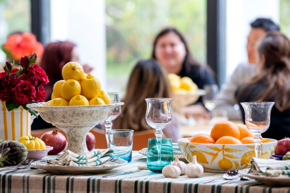 A table with a bowl with oranges and a bowl with lemons, garlic, onions and other vegetables and a vase of Roses and wine and drinking glasses and cutlery, with a table of people in the background
