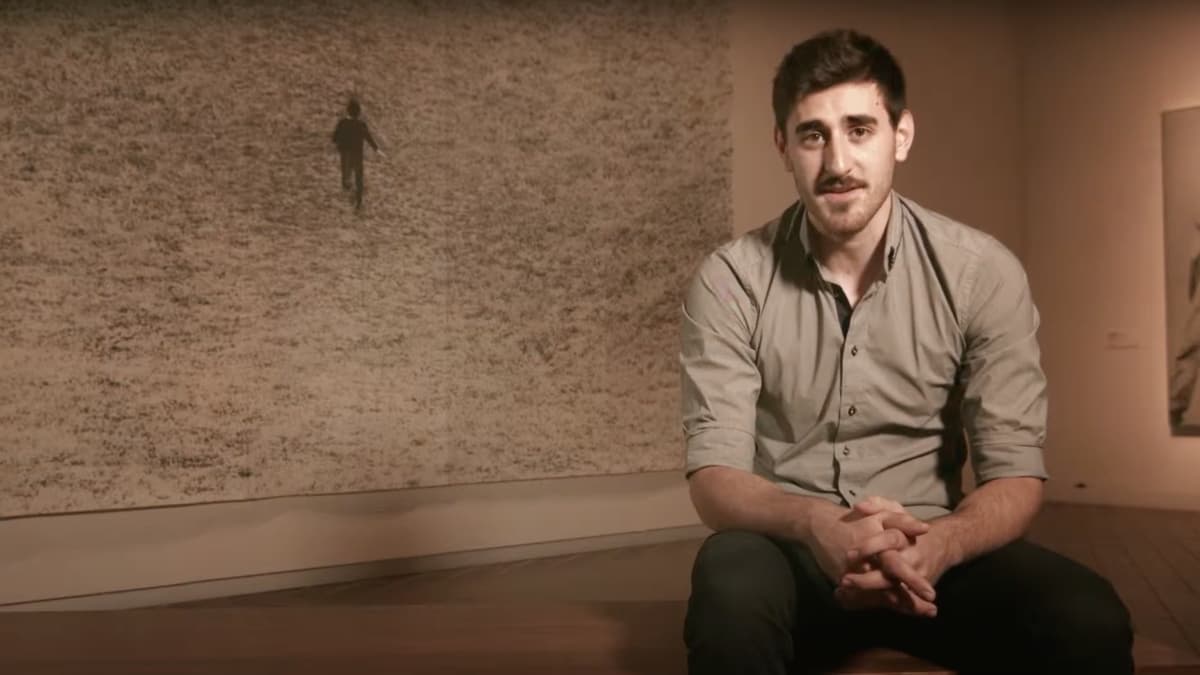 A video still of a man sitting in front of an artwork in a gallery space
