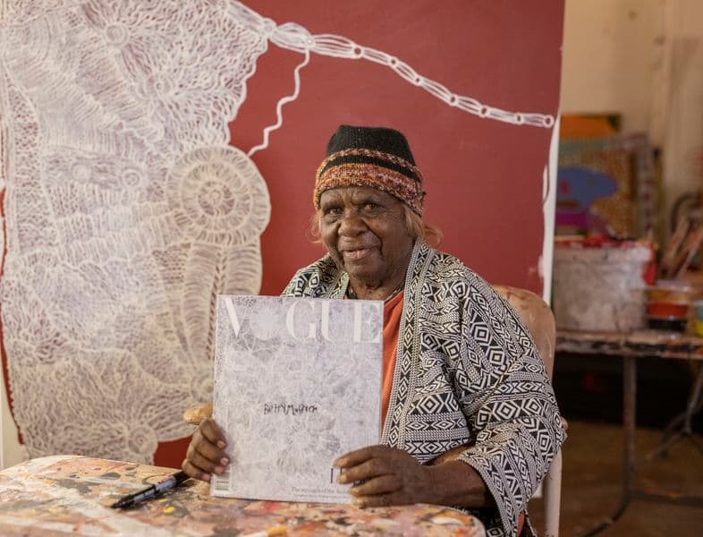 An older woman sits in front of her painting, holding a copy of Vogue magazine that has her artwork on the cover
