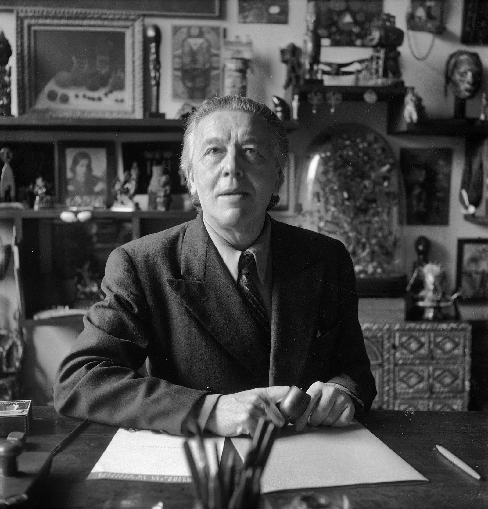 A black and white photo of Andre Breton sitting at a desk, holding a pipe, with a range of objects and artworks on the wall behind him