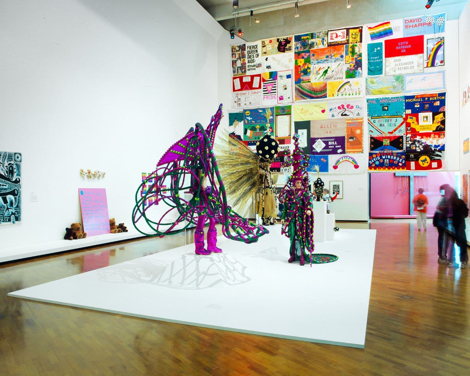 Wide gallery space, white walls featuring a plinth in the centre with bright, glittering mardi gras costumes and behind it a poster wall