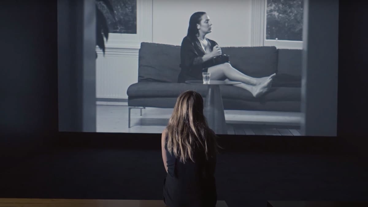 Screen shot of a film still showing a woman from behind, sitting on a bench and watching a large film projection