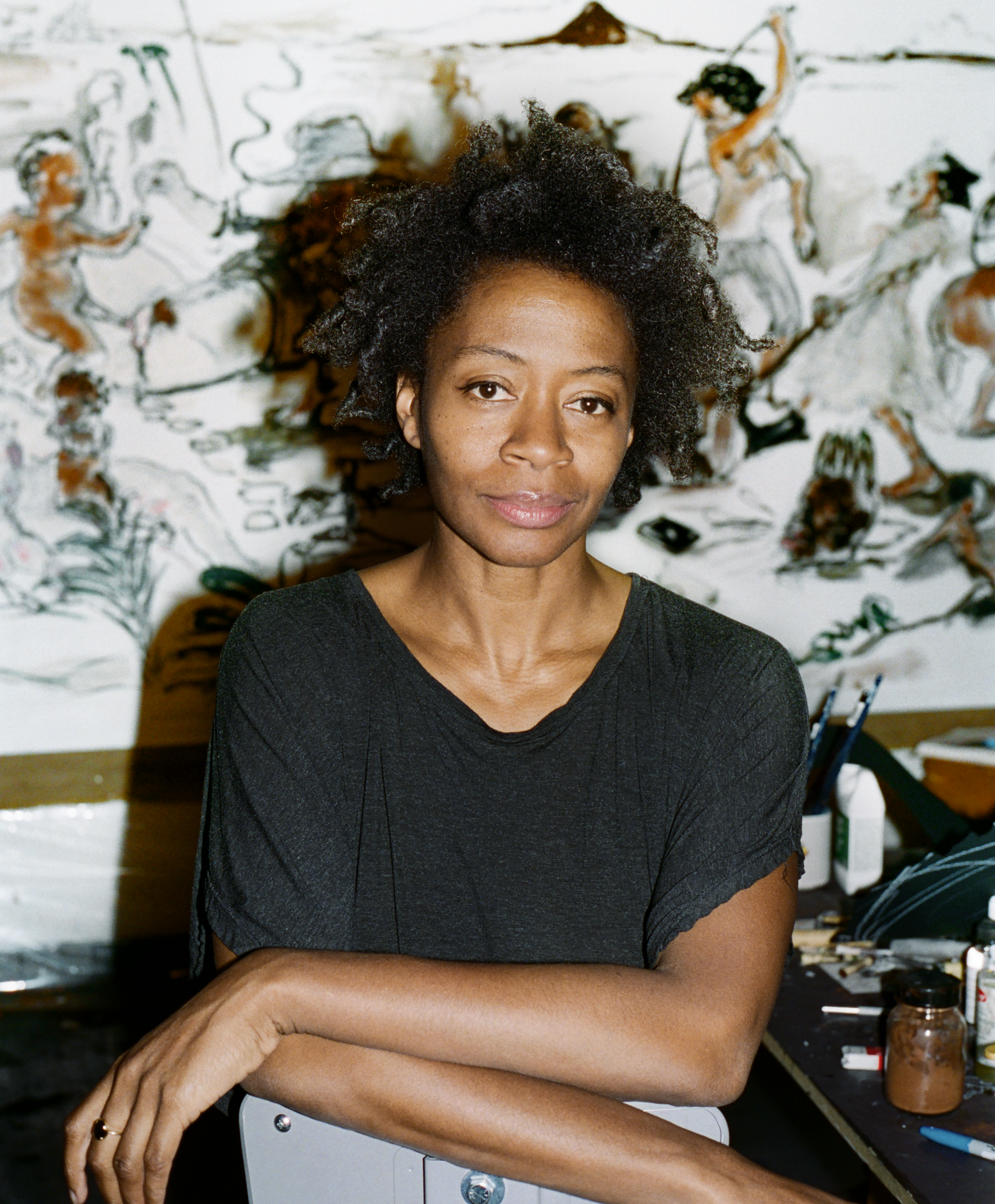 A photographic portrait of a woman artist in a black t-shirt with studio works in the background