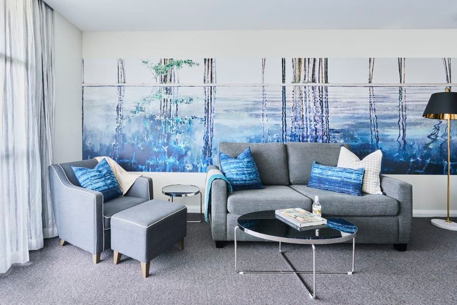 Image of a room with grey couches, blue cushions, and wallpaper of a painting of water and trees