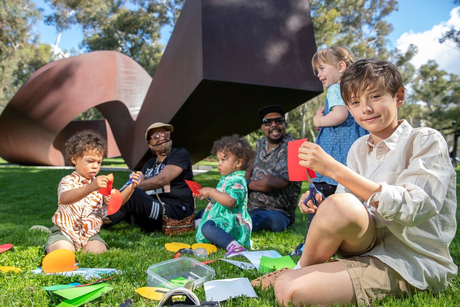 Two adults sit with four children in front of a sculpture in the sculpture garden making art with paper