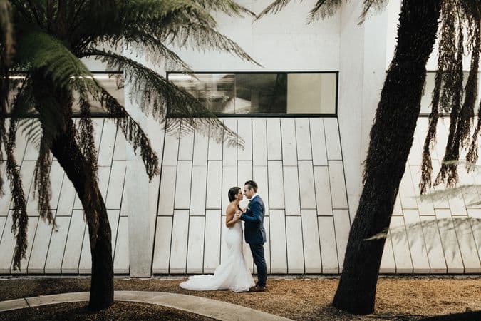 A couple stand in wedding attire, facing each other and holding hands, surrounded by fern trees