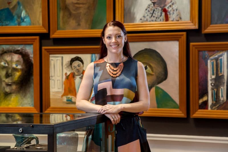Portrait photo of a female with long auburn hair wearing a colourful sleeveless dress, standing in front of a wall of art inside a Gallery