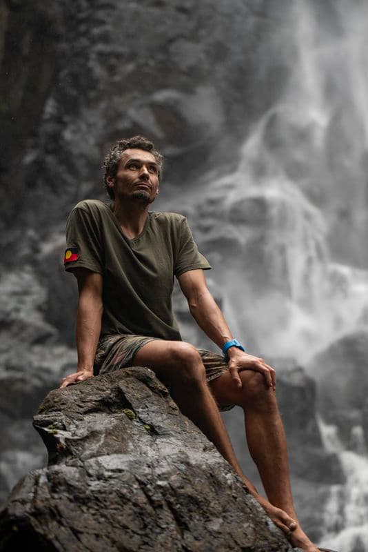 Photograph of man sitting on rock looking up at waterfall