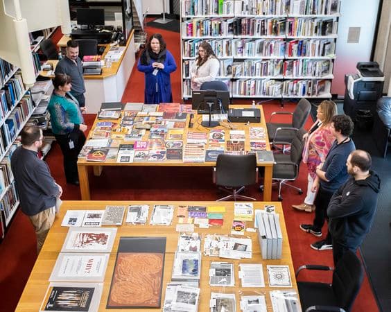 Birds-eye view of two large tables in a library, surrounded by book shelves. Each table has archival material laid out on it, and people are standing around the tables