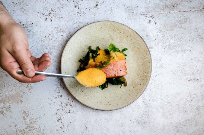 A plate of salmon on a bed of greens, showing a hand holding a spoon with orange foamy liquid pouring over the salmon