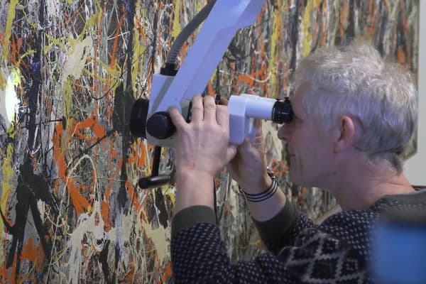 Video still of man looking at painting closely through a microscope.
