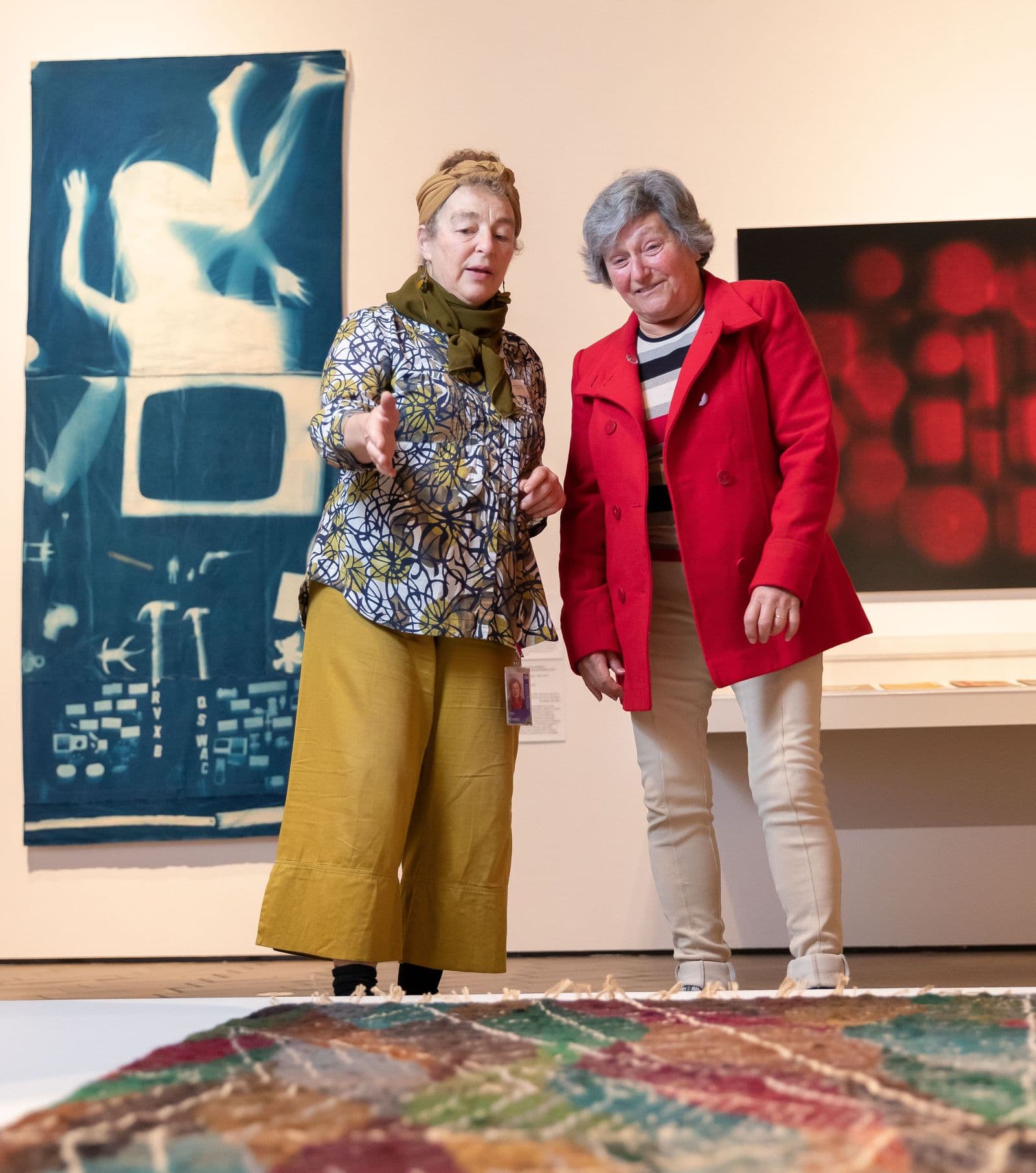In a gallery one woman is pointing and describing a work of art to another woman