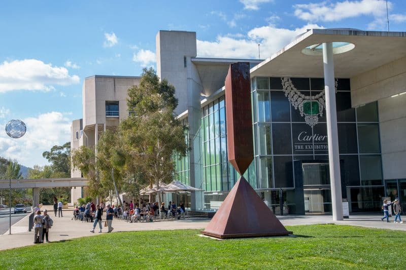 Large sculptures, trees and people at a cafe are in front of a large concrete Brutalist building