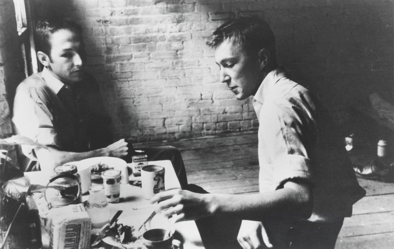 A black and white image of two men sitting around a rectangular table with crockery and food, and a brick wall behind them