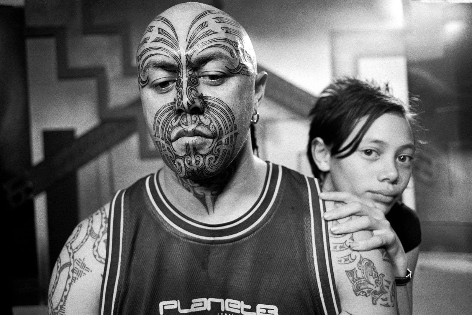 A black and white photo of a man with Moari facial tattoos with a young woman standing behind him, looking over his shoulder