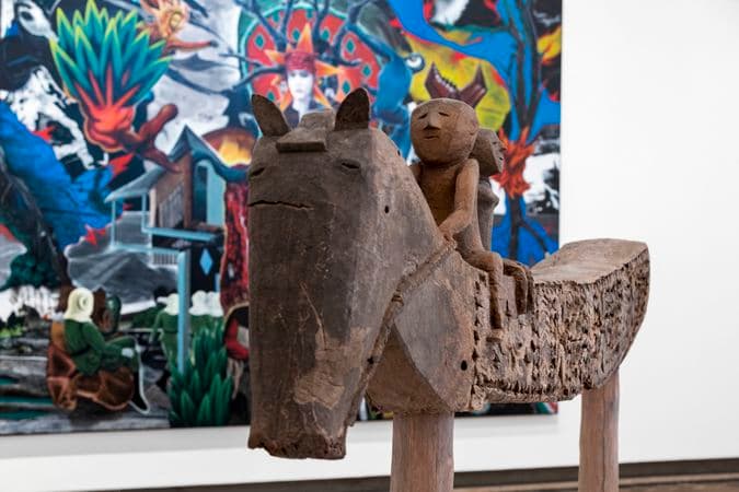 Two figures are seated on a wooden carved horse that is very long in length. Gallery installation photograph.