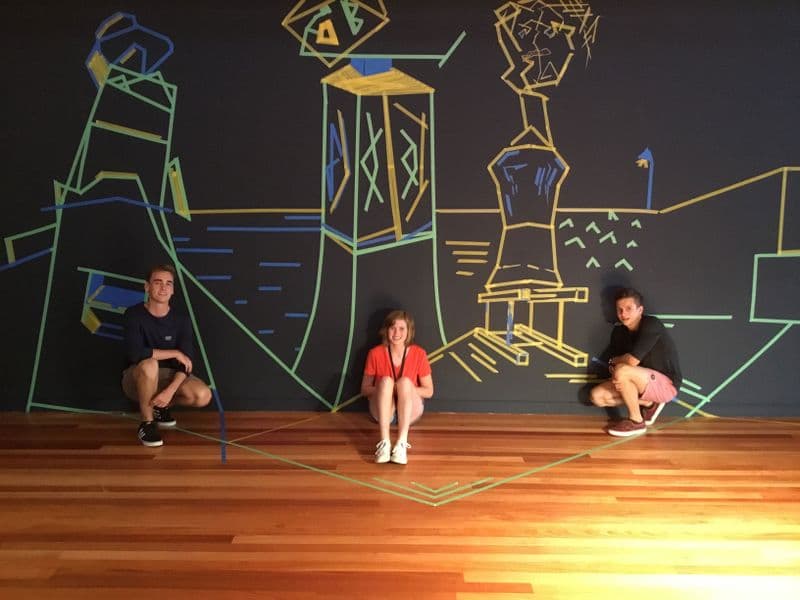three teenagers are sitting in front of a large painted indoor mural on a black wall