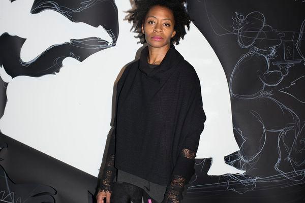 A portrait of the artist Kara Walker - an African American woman standing against a black and white background of her painting. Books and art making materials in the foreground