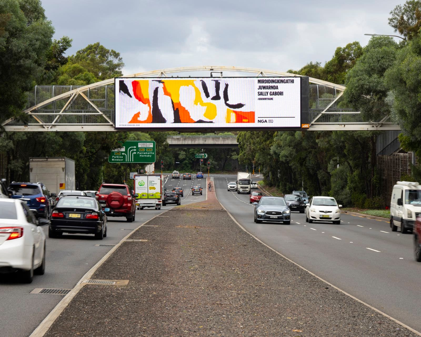 A billboard depicting an indigenous Australian artwork sits above a busy multilane road