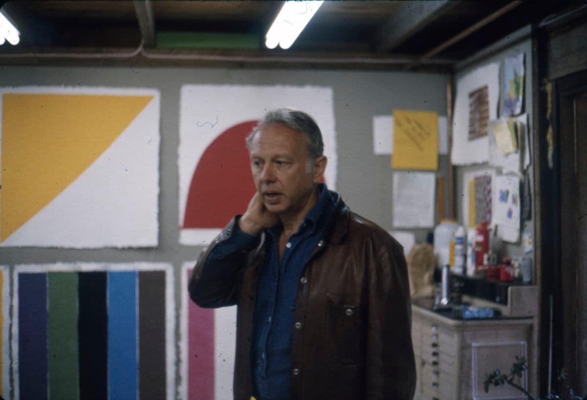 The artist Ellsworth Kelly stands in front of a wall of very colourful artworks made of paper pulp.