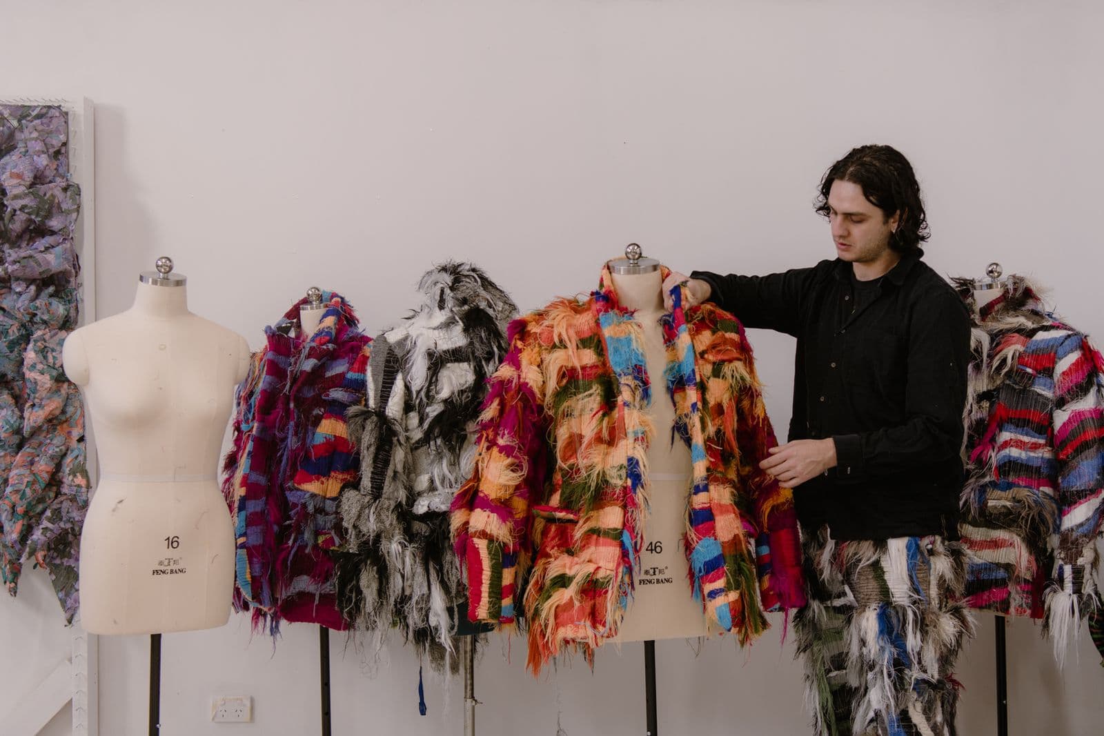 A young person stands in their studio surrounded by colourful items of clothing they have made