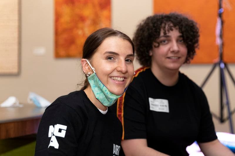 Teenage girl in a Gallery space wearing a mask pulled down off her face, with a teenage boy with dark curly hair in the background.
